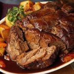 A succulent roast beef slice drenched in glossy rich gravy, served on a white plate with roasted potatoes and a sprinkle of fresh parsley, presented on a wooden surface.