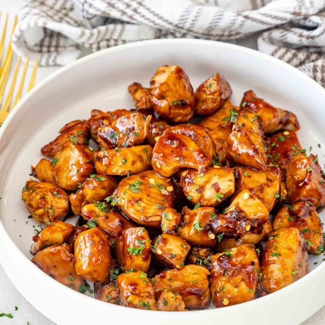 A white bowl filled with tender chicken pieces coated in a glossy honey garlic sauce, garnished with parsley and red chili flakes. A gold fork and a checkered cloth are visible in the background.