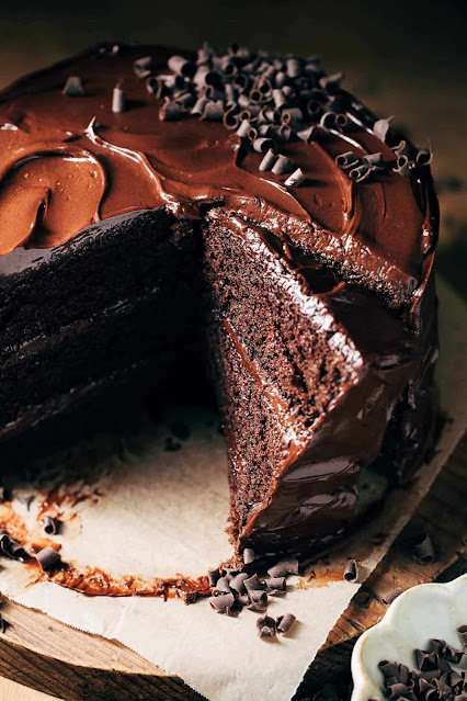 A decadent chocolate cake with a smooth, glossy chocolate frosting, sprinkled with chocolate shavings, and a slice cut out to show its moist, dark interior. Presented on a wooden board with scattered shavings and a small bowl of additional chocolate pieces