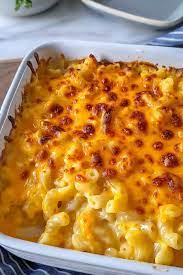 A white casserole dish filled with baked macaroni and cheese, topped with a bubbly, golden-brown cheddar cheese layer. Set on a marble surface with a blue cloth and a small bowl in the background.