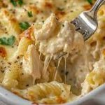 A white dish filled with creamy chicken pasta bake, featuring tender pasta and chicken chunks in a stretchy cheesy sauce, garnished with parsley and black pepper, with a fork lifting a portion.
