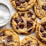 A batch of golden, chewy chocolate chunk cookies sprinkled with coarse sea salt, arranged on parchment paper. A small bowl of sea salt is visible nearby.
