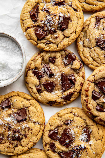 A batch of golden, chewy chocolate chunk cookies sprinkled with coarse sea salt, arranged on parchment paper. A small bowl of sea salt is visible nearby.