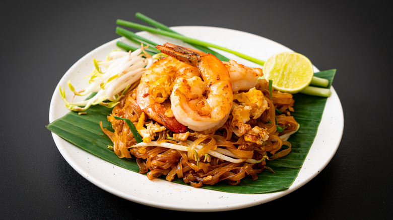 Authentic shrimp Pad Thai on banana leaf with lime wedge and green onions, chopsticks picking up noodles.