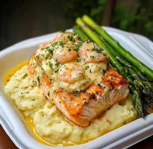 Seared salmon topped with creamy garlic shrimp sauce served over mashed potatoes with roasted asparagus on a white plate.