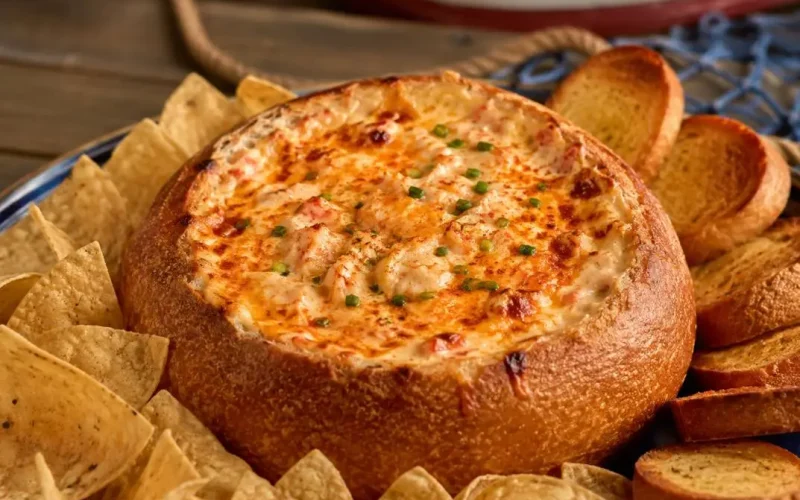 Creamy baked crab dip served in a rustic bread bowl, topped with melted golden cheese and fresh herbs, surrounded by crispy tortilla chips and toasted bread slices.